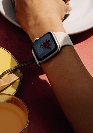 Arm einer Person, die nach etwas zum Frühstücken greift, Apple Watch SE 3 am Handgelenk, Aluminiumgehäuse in Polarstern, Always-On Display, Porträt einer Person auf dem Zifferblatt, Sportarmband in Polarstern